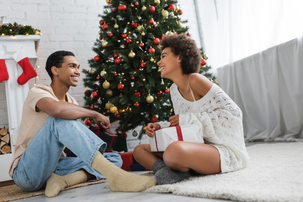 fröhliches afrikanisch-amerikanisches Paar sitzt auf dem Boden neben dem Weihnachtsbaum und schaut einander an  - Foto, Bild