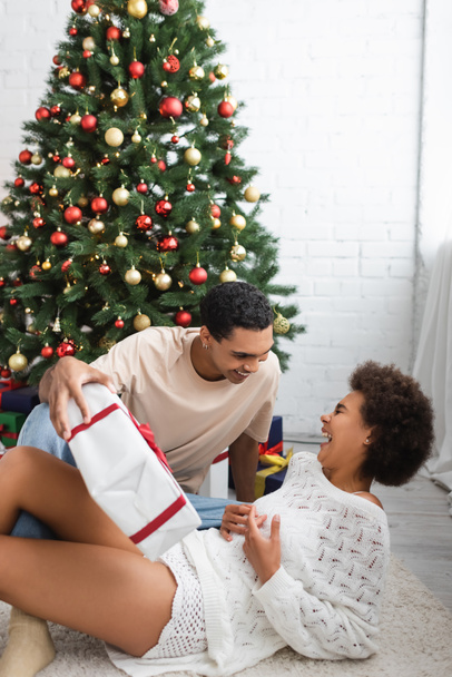 joven afroamericano hombre sosteniendo caja de regalo cerca de árbol de Navidad y novia riendo - Foto, Imagen