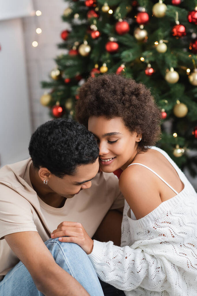 Genç erkek arkadaşının yanında gözleri kapalı gülümseyen seksi Afrikalı Amerikalı kadın ve bulanık Noel ağacı. - Fotoğraf, Görsel