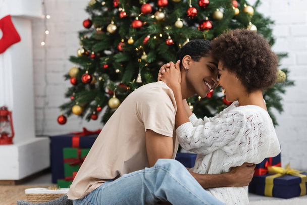 πλαϊνή άποψη των νέων και σέξι Αφροαμερικανό ζευγάρι αγκαλιάζει κοντά χριστουγεννιάτικο δέντρο στο σπίτι - Φωτογραφία, εικόνα
