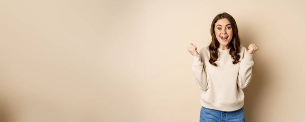 Enthusiastic brunette woman laughing and smiling, cheering, shaking hands and celebrating, triumphing, standing over beige background. - Foto, afbeelding