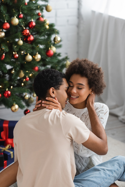 young african american woman with closed eyes hugging boyfriend on christmas day at home - Photo, Image