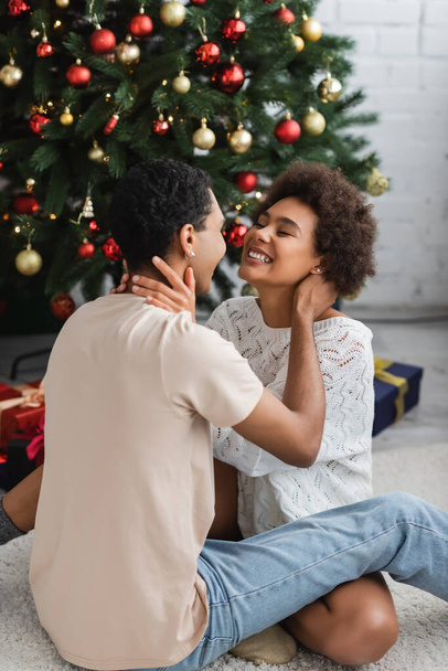 eccitata donna afroamericana che si abbraccia con il fidanzato mentre siede sul pavimento vicino all'albero di Natale - Foto, immagini