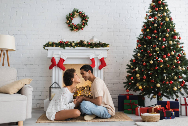 side view of happy african american couple sitting on floor near decorated fireplace and christmas tree - Photo, Image