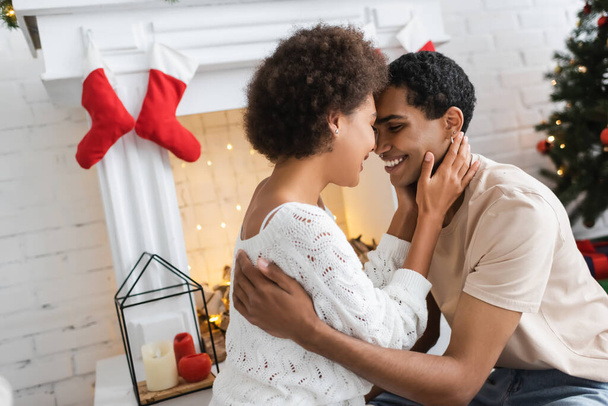 young and happy african american couple embracing near fireplace with christmas decor - Zdjęcie, obraz
