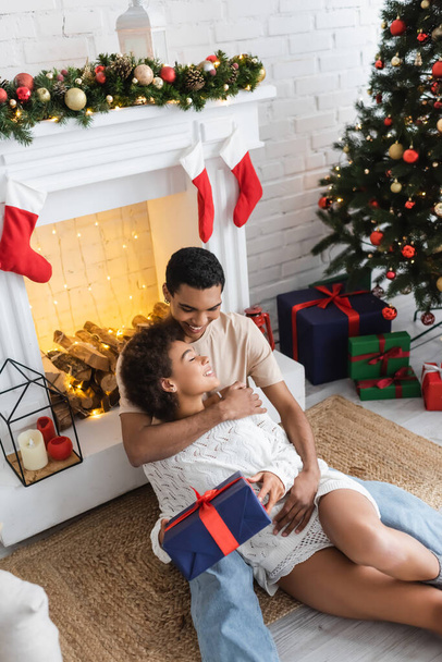 african american man hugging girlfriend sitting with christmas present near decorated fireplace - Photo, Image