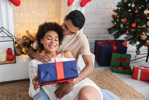 pleased african american woman with gift box sitting near young boyfriend and fireplace at home - Photo, Image