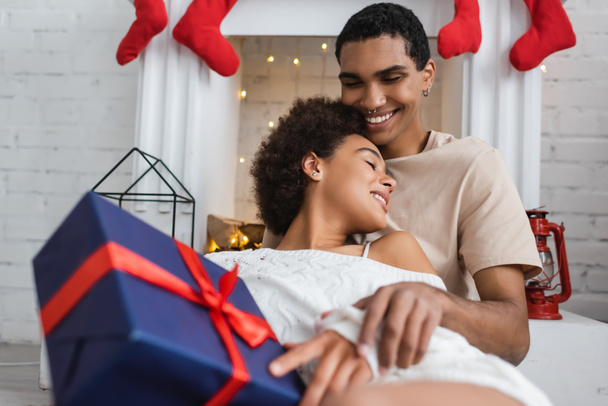 сексуальная женщина с подарком на Рождество рядом с улыбающимся африканским американским парнем, сидящим у камина - Фото, изображение
