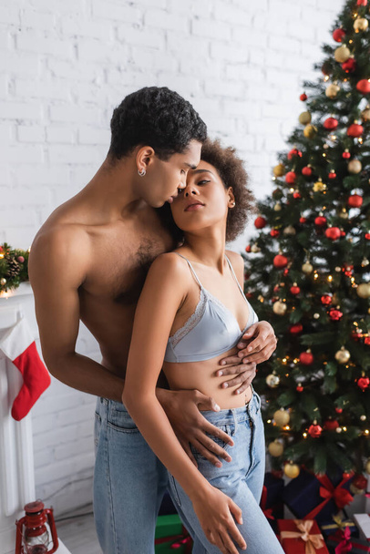 appassionata donna afroamericana guardando l'uomo senza maglietta che la abbraccia vicino all'albero di Natale - Foto, immagini