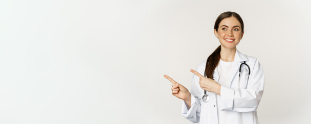 Retrato de una joven doctora sonriente, trabajadora médica, señalando con los dedos hacia la izquierda, mostrando promoción clínica, logotipo o pancarta, de pie sobre fondo blanco. - Foto, Imagen
