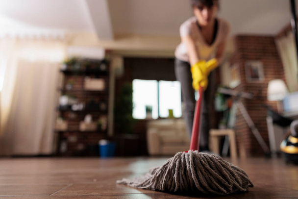 Housemaid πάτωμα καθαρισμού με σφουγγαρίστρα φορώντας γάντια καθαρισμού, φινίρισμα διαμέρισμα καθαρισμού για πελάτη. Γυναίκα οικιακή υπάλληλος κάνει τη δουλειά της με αφοσίωση, συντήρηση σπιτιού. - Φωτογραφία, εικόνα