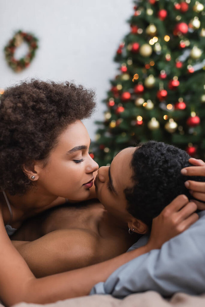curly and sexy african american woman kissing boyfriend near blurred christmas tree at home - Foto, afbeelding