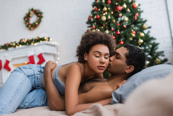 appassionata donna afroamericana ad occhi chiusi che abbraccia l'uomo in camera da letto con decorazioni natalizie sfocate - Foto, immagini
