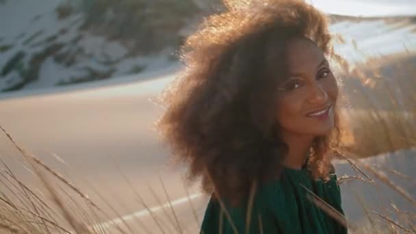 Portrait de femme aux cheveux noirs heureuse dans le désert d'été avec un maquillage moderne. Belle fille afro-américaine souriante regardant de près la caméra des dunes de sable. Brune bouclée joyeuse assise près de l'herbe sèche. - Séquence, vidéo