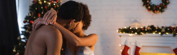 sexy afrykański amerykański para przytulanie i całowanie w pobliżu rozmazane świąteczne dekoracje, sztandar - Zdjęcie, obraz