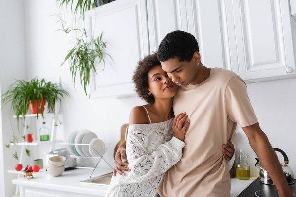 sexy african american woman in white knitted sweater smiling near young boyfriend in kitchen - Foto, immagini