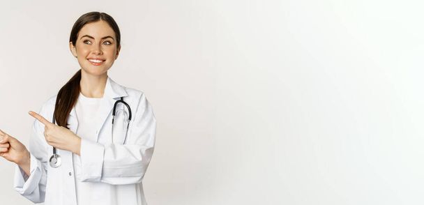 Πορτρέτο της χαμογελαστή νεαρή γυναίκα γιατρός, ιατρική περίθαλψη εργαζόμενος, δείχνοντας τα δάχτυλα αριστερά, δείχνει κλινική promo, λογότυπο ή πανό, στέκεται πάνω από λευκό φόντο. - Φωτογραφία, εικόνα