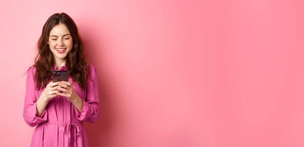 Mujer joven de compras, charlando en el teléfono móvil, sonriendo mientras lee la pantalla del teléfono inteligente, de pie sobre el fondo rosa. Copiar espacio - Foto, Imagen