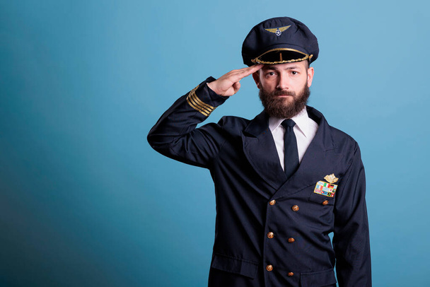 Σοβαρός πιλότος αεροπλάνου χαιρετά, φοράει στολή και καπέλο μπροστά πορτρέτο άποψη, καπετάνιος αεροπλάνο κοιτάζοντας κάμερα. Αεροπορική ακαδημία αεροπόρος με σήμα φτερά αεροπορικής εταιρείας στο σακάκι - Φωτογραφία, εικόνα