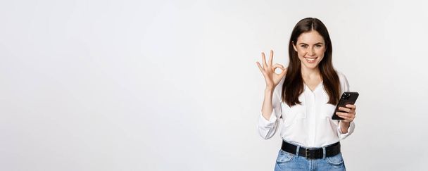 Smiling woman showing okay, ok sign while using mobile phone, approve app on smartphone, excellent service, online store, standing over white background. - 写真・画像