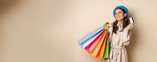 Portrat des trendigen femininen Mädchens posiert mit Einkaufstaschen aus dem Geschäft und Kreditkarte, kontaktlos bezahlen, mit Rabatt kaufen, beiger Hintergrund. - Foto, Bild