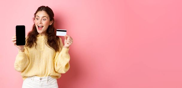 Technologie a online nakupování. Vzrušený atraktivní dívka ukazující displej smartphone, plastové kreditní karty, vypadají ohromeně na telefonu, stojí na růžovém pozadí. - Fotografie, Obrázek