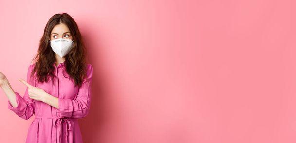 Ковид-19, концепция карантина и социального дистанцирования. Любопытная стильная женщина в респираторе, смотрящая и указывая пальцами в сторону, показывая логотип промо-текста, стоя на розовом фоне. - Фото, изображение
