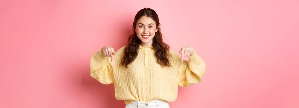Portrait of young attractive female model pointing down at copy space, showing promotional text or logo, standing against pink background. - Photo, Image