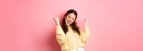 Girl screams with joy and fist pump, say yes, achieve goal or success, celebrating achievement, triumphing and winning, standing over pink background. - Foto, immagini