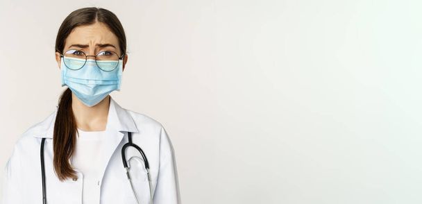 Sad and gloomy young woman doctor in medical face mask, feeling upset or concerned, frowning, standing over white background. Copy space - Foto, afbeelding