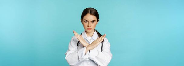 Портрет лікаря-жінки, який показує хрест, припинити забороняти жест, заборонити смоктати, стоячи в білому пальто на бірюзовому фоні
. - Фото, зображення