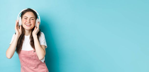 Симпатична біла дівчина у весняному вбранні, слухаючи музику в навушниках, усміхаючись задоволена камерою, стоїть над синім тлом
. - Фото, зображення