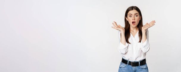 Portrait of concerned female model looking shocked, surprised face reaction, standing in shirt over white background. Copy space - Фото, зображення