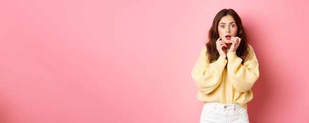 Portrait of scared and shocked brunette girl, gasping and holding hands near lips, staring frightened at camera, standing startled against pink background. - Photo, Image