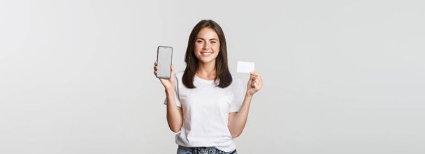 Aantrekkelijk lachend meisje op zoek tevreden en tonen credit card, mobiele telefoon scherm. - Foto, afbeelding