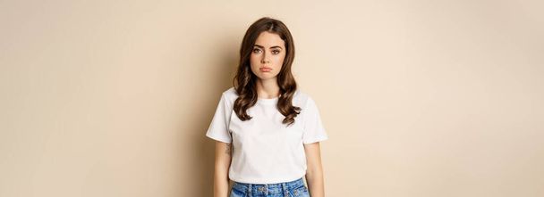 Sad and tired brunette woman looking drained and unamused, standing with pokerface against beige background. - Fotó, kép