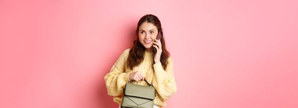 Menina glamour jovem segurando sua bolsa, falando no smartphone e sorrindo, tendo uma conversa casual no telefone, chamando alguém, de pé sobre fundo rosa. - Foto, Imagem