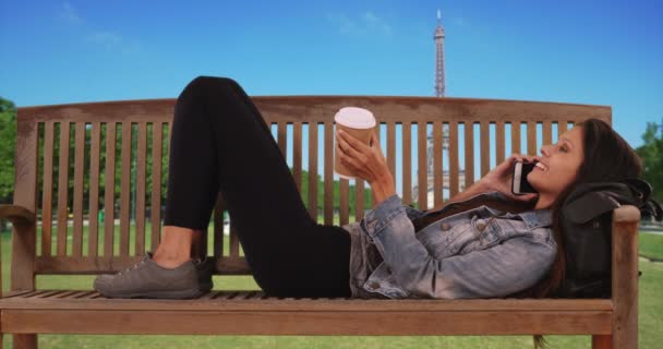 Tisíciletá žena ležící na lavičce v parku poblíž Eiffelovy věže, zatímco telefonovala s kamarádkou. Kavkazská turistka ve Francii leží s kávou a chatuje na mobilu. 4k - Záběry, video