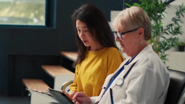 Yaşlı doktor ve Asyalı kadın check-up randevusu yapıyor, tıbbi rapor yazıyor ve dijital tableti kontrol ediyor. Tesis lobisinde konsültasyondan önce sigorta formu doldurmak. - Video, Çekim