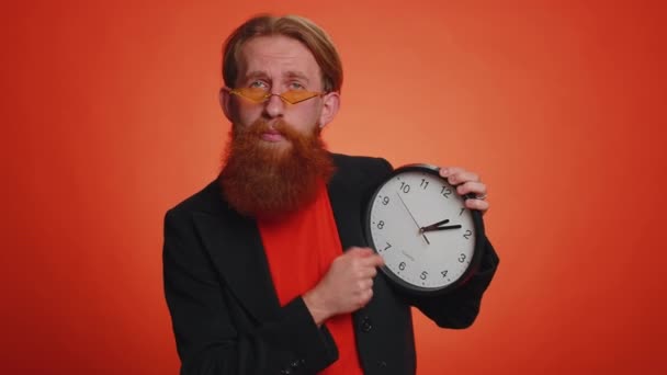 It is your time. Portrait of bearded young man in jacket showing time on clock watch, ok, thumb up, approve, pointing finger at camera. Adult guy indoor studio shot isolated alone on orange background - Footage, Video