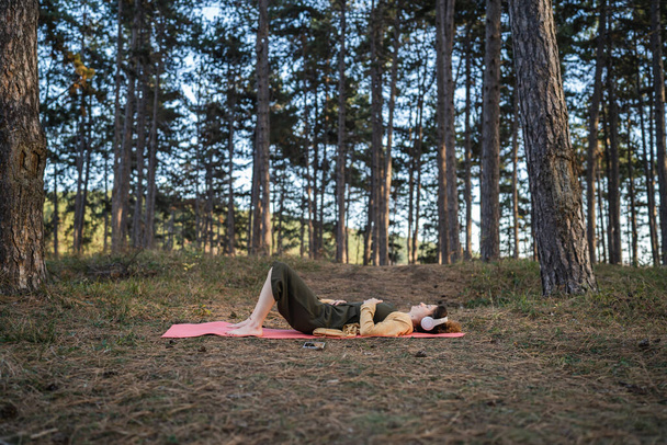 Genç bir kadın beyaz kadın parkta ya da ormanda tek başına uzanıyor. Kulaklıklarıyla meditasyonlara rehberlik ediyor. Kişisel bakım dengesi ve tezahür uygulamaları. - Fotoğraf, Görsel