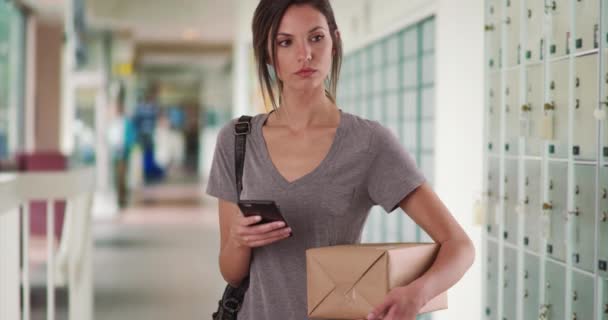 Kobieta korzystająca z telefonu komórkowego podczas trzymania przesyłki pocztowej przez szafki w korytarzu poczty. Millennial kobieta za pomocą aplikacji smartfon do śledzenia pakietu podczas pobytu na poczcie. 4k - Materiał filmowy, wideo