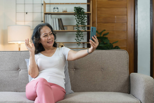 Asian elderly woman taking selfie on mobile phone while sitting on sofa and smiling. - Zdjęcie, obraz