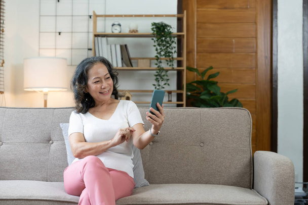 Smiling senior lady using her smartphone, she is connecting online and using apps at cozy home. - 写真・画像