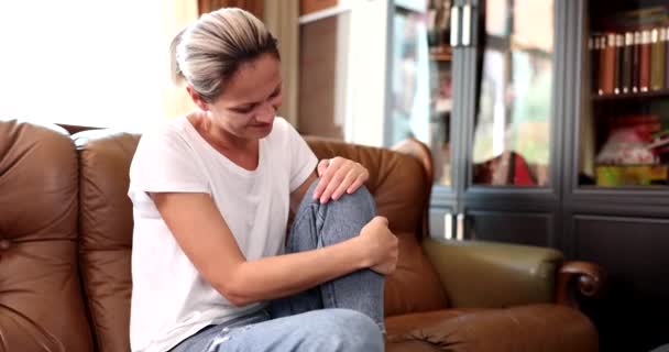 Nő ül a kanapén a nappaliban, és szenved a fájdalom a térd és a láb Masszázs, izomhúzódás és ín összehúzódás - Felvétel, videó