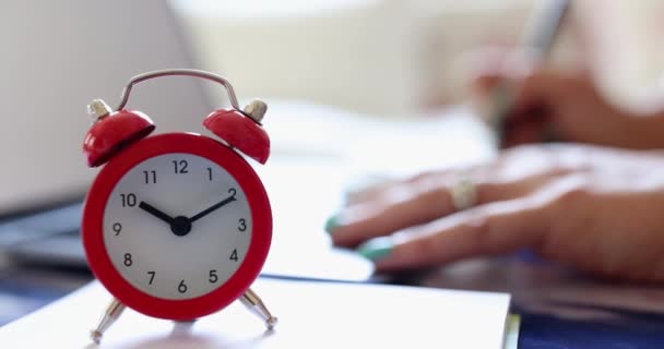 目覚まし時計を持つ女性は、ノートパソコンの前のテーブルに座って、ワークブックに書き込みます。締め切り時間管理オンライン学習時間 - 映像、動画