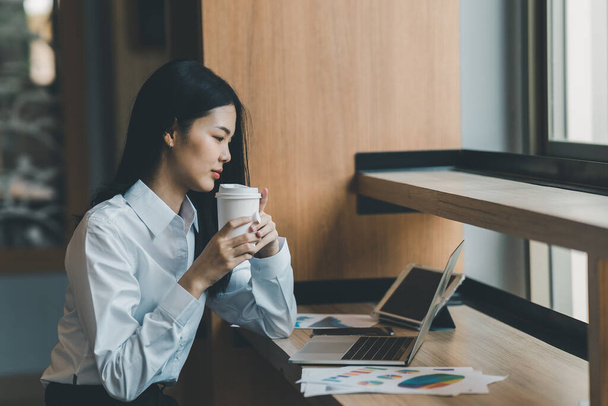 Азиатская фрилансерша улыбается, держа чашку горячего кофе и работая за ноутбуком на деревянном столе в кафе. Предпринимательница, работающая в кофейне. Работа в любой точке мира - Фото, изображение