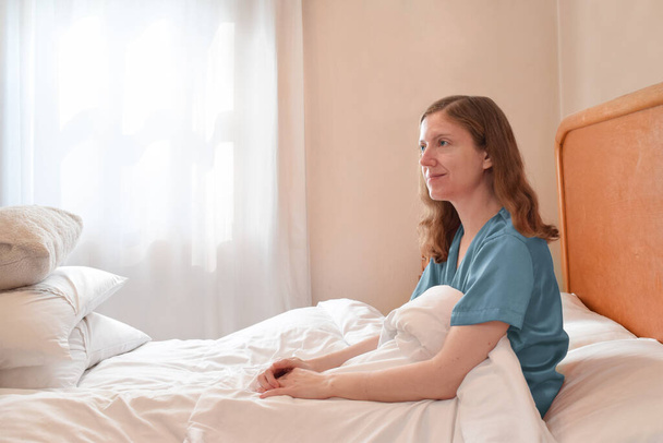 Mulher caucasiana de pijama de seda azul contemplando o dia enquanto sentada na cama. Muito espaço de cópia. - Foto, Imagem