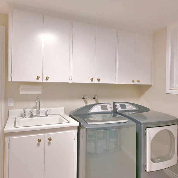 Plac Wnętrze pralni z otwartymi białymi drzwiami i widokiem na korytarz. Przy umywalce pod szafkami ściennymi obok przesuwnych okien po lewej stronie znajdują się pralki i suszarki.. - Zdjęcie, obraz