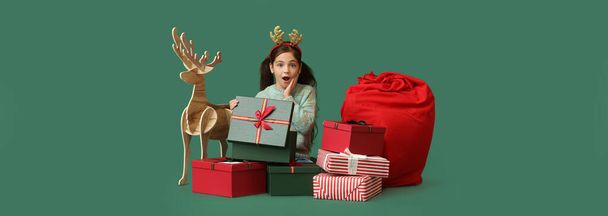 Surprised little girl with wooden reindeer, Santa bag and Christmas gifts on green background - Foto, imagen